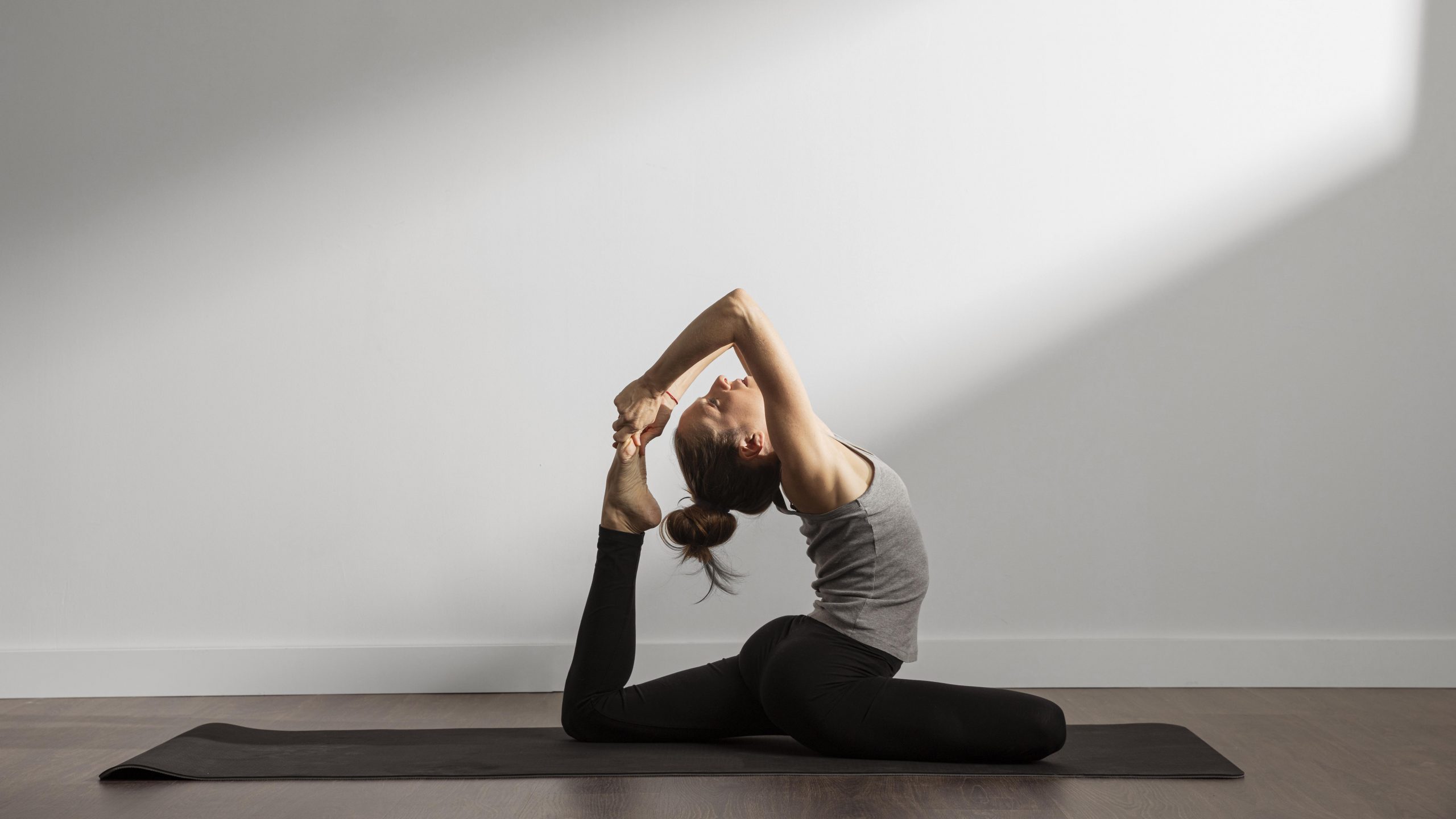 Frau in fortgeschrittener Yoga-Rückbeuge auf einer Matte in einem hellen Raum. Sie hält ihren Fuß mit beiden Händen hinter dem Kopf in der Königstauben-Pose.